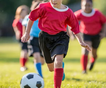 Children playing soccer on a field, with one child in a red jersey running towards the ball.