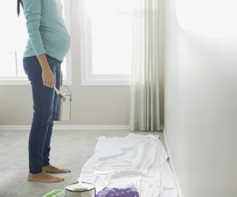 Pregnant woman holding paint can and brush, standing in a bright room with drop cloth on the floor.