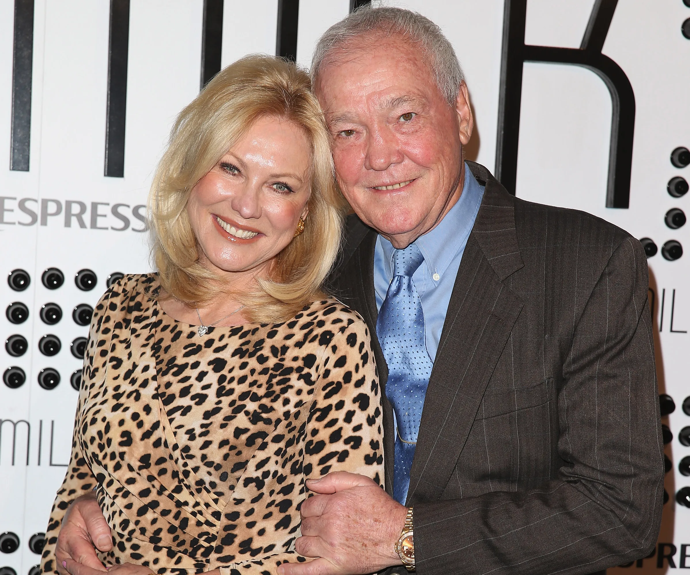 Couple smiling together; woman in leopard print dress, man in suit with blue tie, posing at an event backdrop.