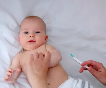 A baby lying on a bed, with an adult hand holding a syringe near the baby, implying vaccination.