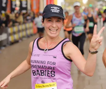 Smiling woman running in charity race, wearing a pink "Running for Premature Babies" shirt and cap, flashing a peace sign.