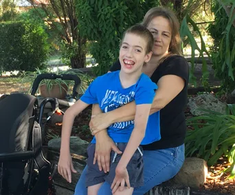 Woman holding a smiling boy in a blue shirt sitting on a bench, with a wheelchair nearby in a garden setting.