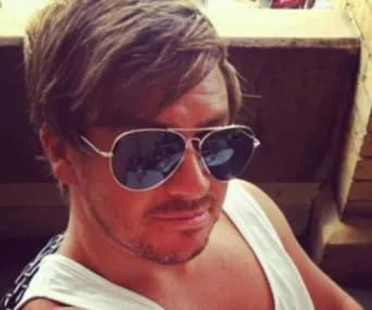 Man wearing sunglasses and a sleeveless shirt, seated outdoors.