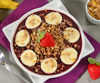 Acai bowl topped with banana slices, granola, and a strawberry on a purple napkin.