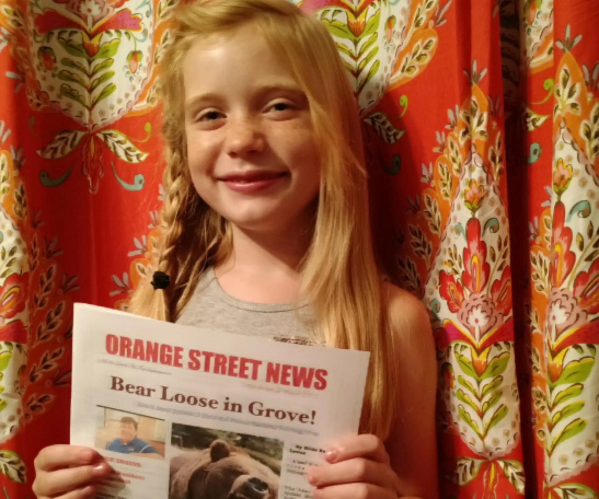 Young girl smiling, holding an "Orange Street News" paper with "Bear Loose in Grove!" headline against a colorful curtain backdrop.