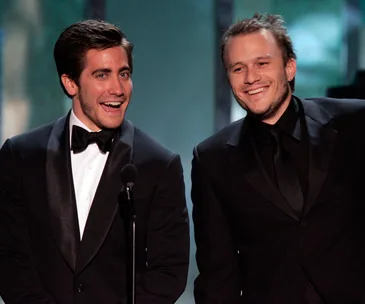 Two men in tuxedos smiling while standing at a microphone at an event.