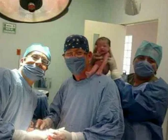 Doctors in surgical attire pose for a selfie holding a newborn baby in an operating room.