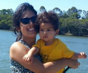 A woman holding a young child near a body of water with trees in the background.