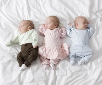 Three newborn triplets sleeping on a white blanket, dressed in pastel outfits.