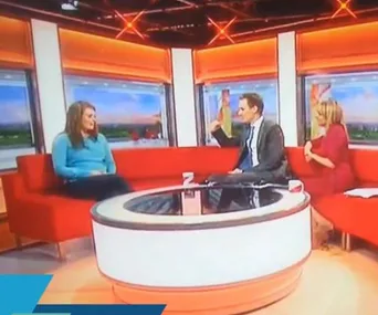 Chat show hosts and guest talking on a red sofa set with orange-lit backdrop and round table in a TV studio.