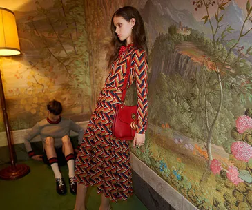 Model in a long, colorful patterned dress, leaning against a wall with floral wallpaper, holding a red handbag.