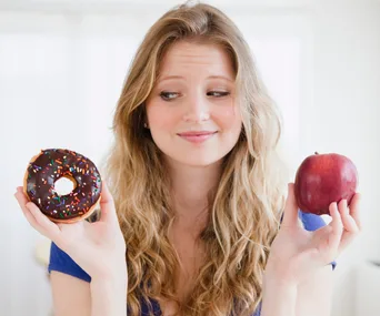 Woman choosing between a chocolate donut with sprinkles and a red apple, looking thoughtful.