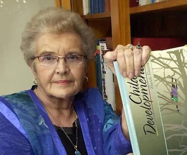 An elderly woman holding a book titled "Child Development" in front of a bookshelf.