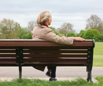 Elderly woman sitting on park bench, facing greenery, overcast sky.
