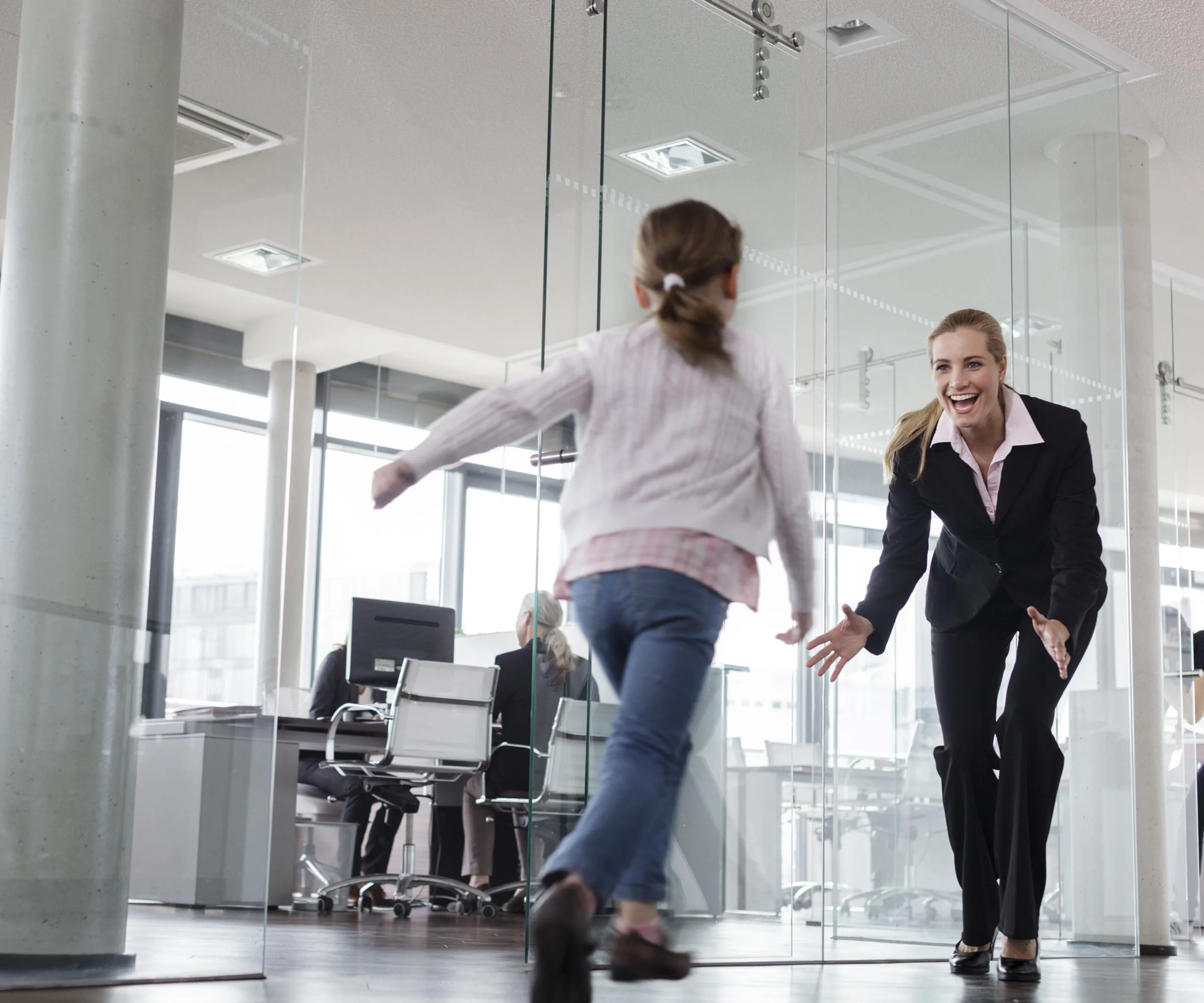 A businesswoman happily greets a young girl running towards her in a modern office setting.