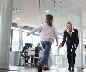 A businesswoman happily greets a young girl running towards her in a modern office setting.