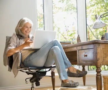 Senior woman smiling while using a laptop at a wooden desk in a bright room.