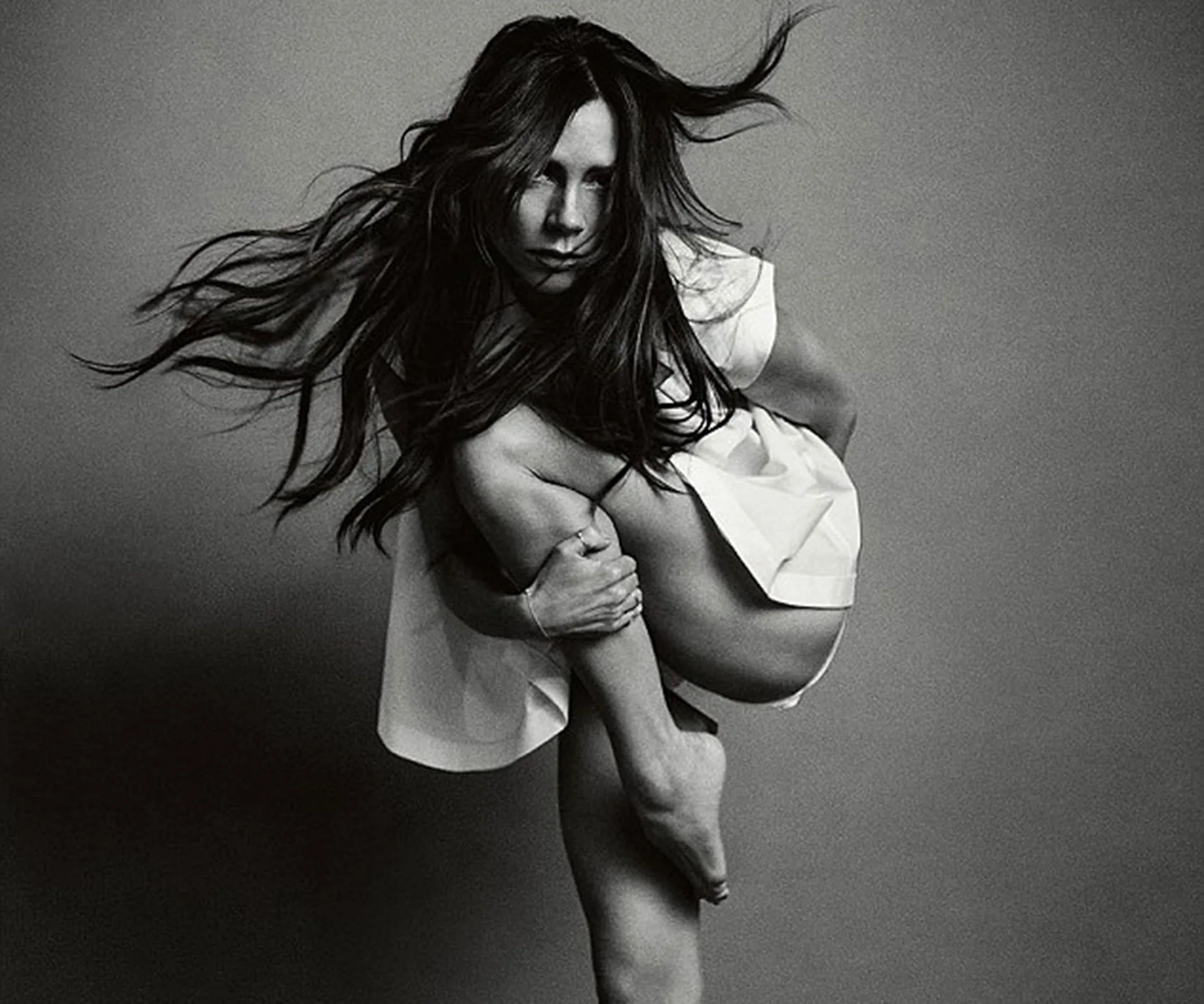 A woman in a dynamic pose with flowing hair and a draped white garment against a grey backdrop.