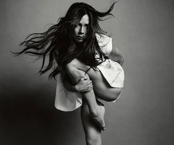 A woman in a dynamic pose with flowing hair and a draped white garment against a grey backdrop.