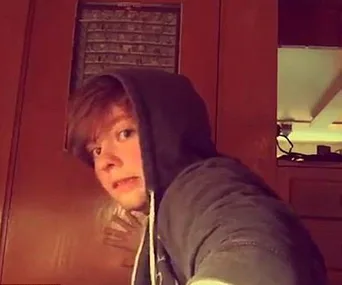 Young person in a hoodie looking at the camera with a surprised expression inside a dimly lit room.