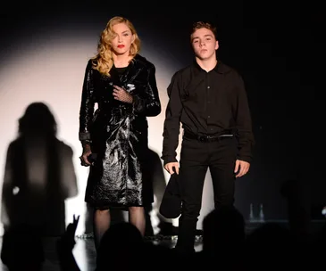 Madonna and a young man stand side by side on stage, both dressed in black, with a dramatic shadow cast behind them.