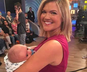 A woman smiling while holding a baby wrapped in a star-patterned blanket in a studio setting with an audience behind.