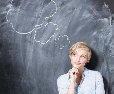 A woman with short blonde hair pensively looks up at drawn thought bubbles on a chalkboard.