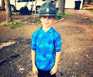 Young boy in blue shirt and grey hat, smiling outdoors, with trees and parked vehicle in background.