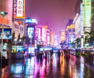 Neon-lit street in Shanghai at night with blurred pedestrians and colorful reflections on wet pavement.