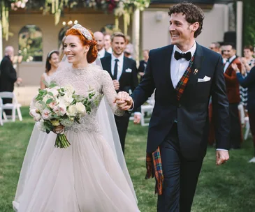 Bride and groom joyfully walk hand in hand through a garden, she holds a bouquet of white flowers, surrounded by guests.