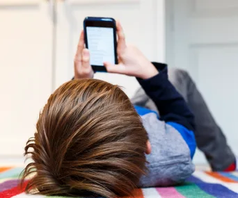 A child lying on a colorful blanket, using a smartphone, with the screen displaying text.