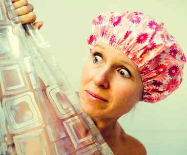 Woman wearing a floral shower cap, peeking from behind a patterned shower curtain with a surprised expression.
