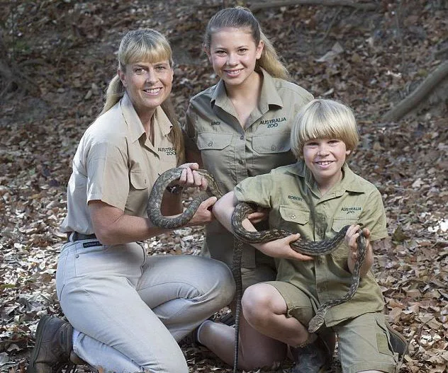 A family in khaki uniforms holding a snake in a wooded area, smiling and crouching together.