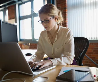 Office worker using a laptop at a desk with a notebook and tablet on a sunny day.