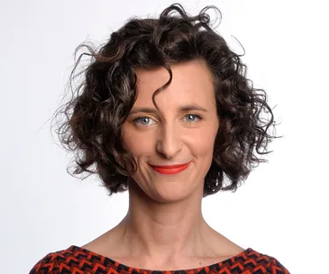 A woman with curly hair smiles at the camera, wearing a patterned red and black top.