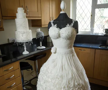A wedding dress cake designed to resemble a bridal gown with intricate lace, placed on a stand in a kitchen setting.