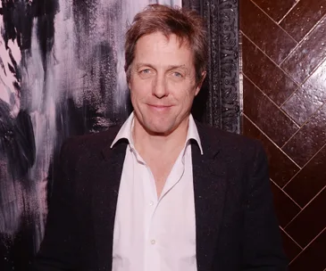 A person smiling, wearing a white shirt and dark jacket, standing in front of an abstract dark and textured background.