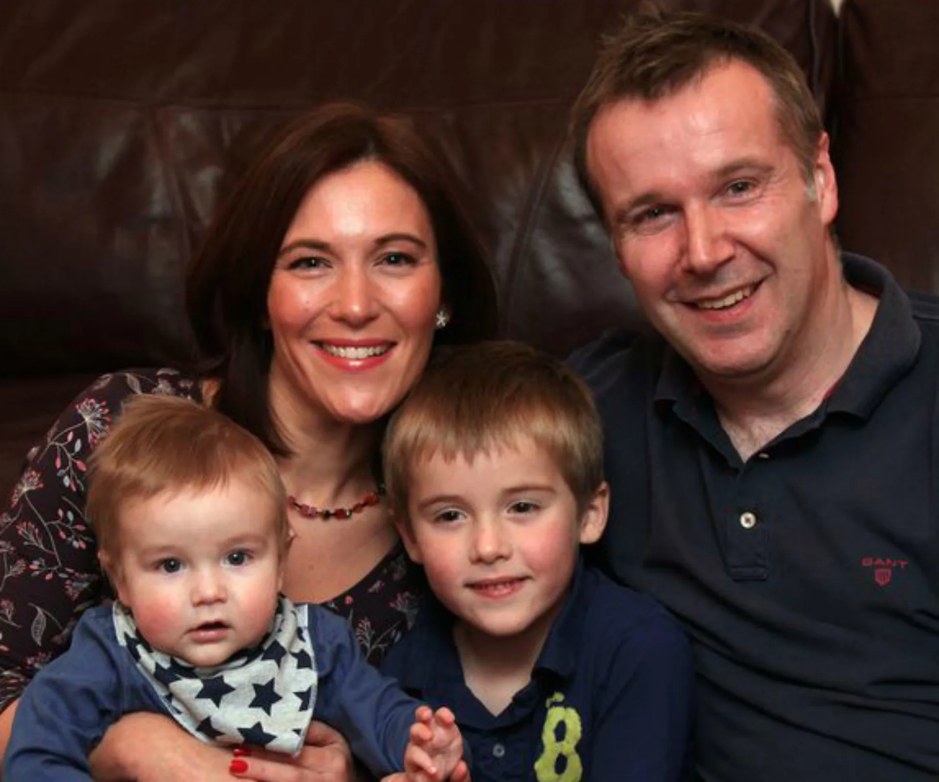 A smiling family: mother, father, young boy, and baby sitting together on a sofa.