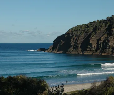 Beach with waves, a rocky cliff, and a few people in the water. A person stands on the sandy shore.