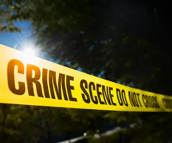 Yellow crime scene tape reading "CRIME SCENE DO NOT CROSS" with sunlight and trees in the background.
