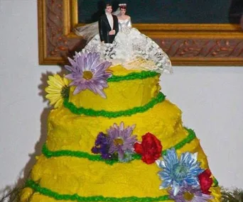 Yellow wedding cake with green icing, adorned with colorful flowers and topped with a bride and groom figurine.
