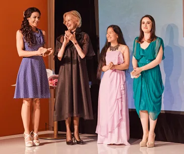 Women in elegant dresses stand together on stage, smiling and clapping, at a formal event against a simple backdrop.