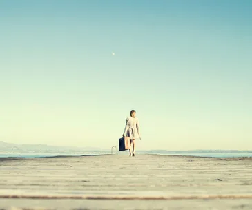 A woman in a coat walks alone on a boardwalk with a suitcase, against a clear sky and distant hills.