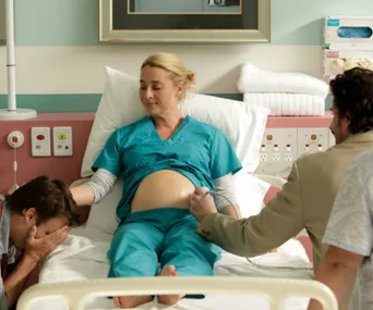 A pregnant woman in hospital scrubs lies on a bed, with two men attending to her, one wiping his face.