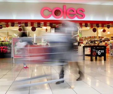 Blurred shoppers with trolleys move past the entrance of a Coles supermarket, featuring promotional signs and bright lighting.
