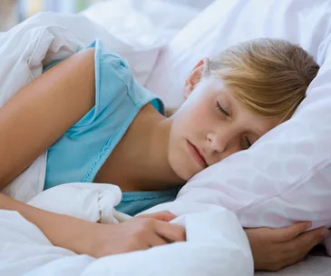 A child in a blue shirt sleeps peacefully on a white and pink pillow, emphasizing the benefits of a consistent bedtime.