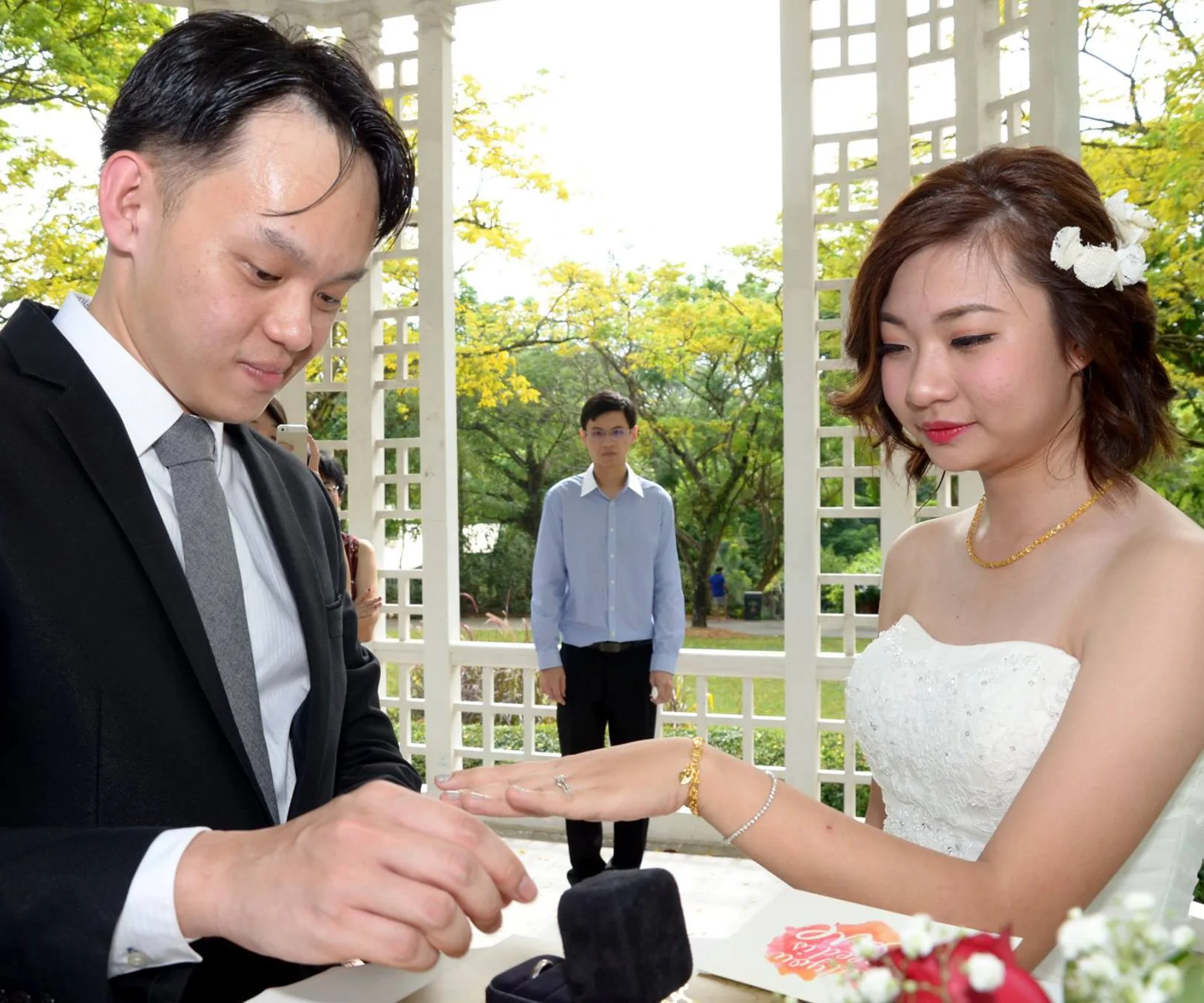 Groom places ring on bride's finger while another man stands in the background at an outdoor wedding ceremony.