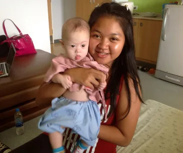 A woman smiling while holding a baby in a cozy room, with a pink bag and laptop nearby.