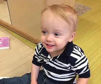 Smiling toddler with blonde hair and a striped shirt sits on the floor near a toy book.