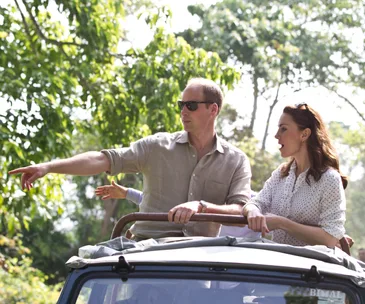 Royal couple on safari, pointing from an open-top vehicle in a lush, green environment.